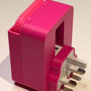 Puede incluir: Una estaci&oacute;n de carga rosa brillante, impresa en 3D, con un enchufe del Reino Unido. La estaci&oacute;n tiene un compartimento para un reloj y una ranura para el enchufe. El dise&ntilde;o es moderno y funcional, con una est&eacute;tica minimalista.