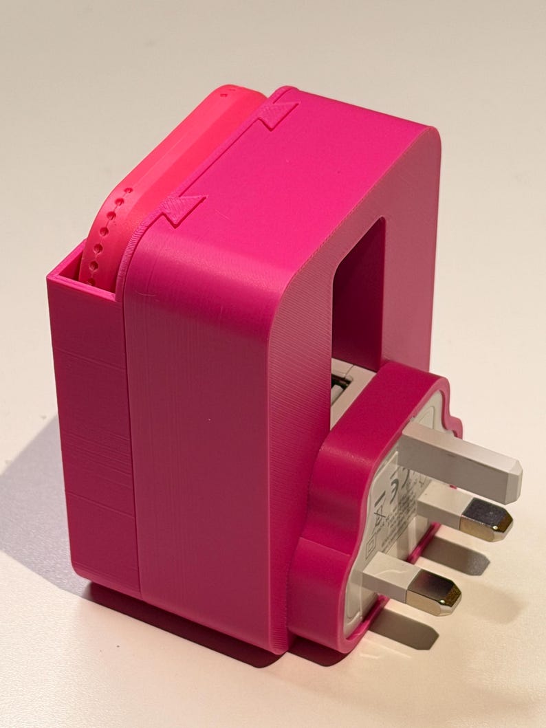 Puede incluir: Una estaci&oacute;n de carga rosa brillante, impresa en 3D, con un enchufe del Reino Unido. La estaci&oacute;n tiene un compartimento para un reloj y una ranura para el enchufe. El dise&ntilde;o es moderno y funcional, con una est&eacute;tica minimalista.