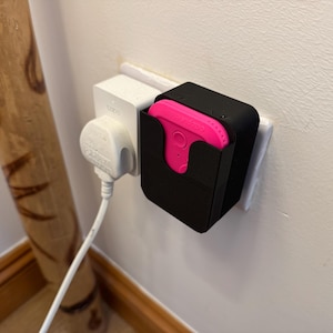 May include: A black electrical outlet adapter with a pink device plugged into it. The adapter is mounted on a white wall.
