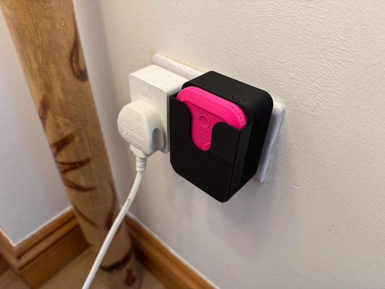 May include: A black electrical outlet adapter with a pink device plugged into it. The adapter is mounted on a white wall.
