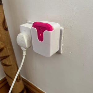 May include: A white plastic wall mount with a pink smart home device plugged in. The device has a red light on the front.