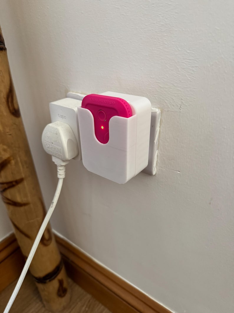 May include: A white plastic wall mount with a pink smart home device plugged in. The device has a red light on the front.