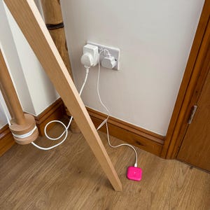 May include: A white electrical outlet with two plugs inserted and a pink square-shaped device plugged into a white cord on a wooden floor.