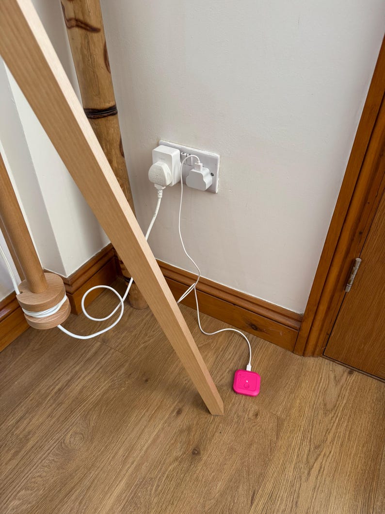 May include: A white electrical outlet with two plugs inserted and a pink square-shaped device plugged into a white cord on a wooden floor.