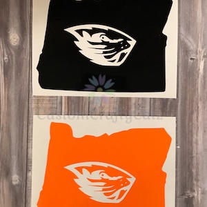 May include: Two vinyl decals of the state of Oregon with a white beaver head inside. The decals are on a white background. One decal is black and the other is orange.