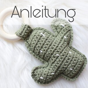 May include: A green crocheted cactus toy with white stitching attached to a wooden teething ring. The toy is on a white fluffy surface. The text "Anleitung" is visible above the toy.