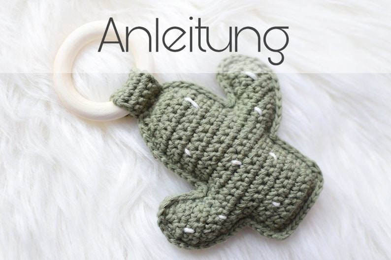 May include: A green crocheted cactus toy with white stitching attached to a wooden teething ring. The toy is on a white fluffy surface. The text "Anleitung" is visible above the toy.