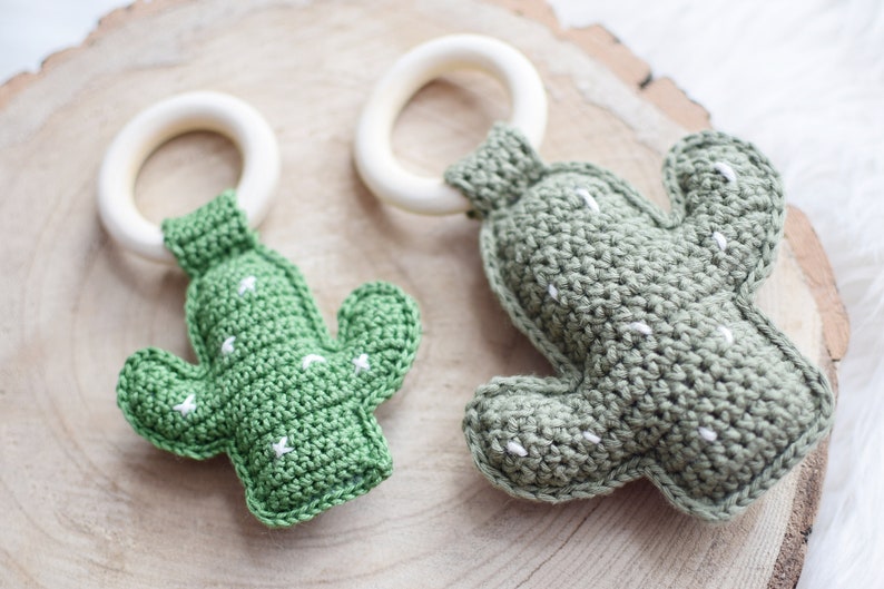 May include: Two crocheted cactus toys with wooden teething rings. The cactus toys are green with white stitching and the rings are natural wood.