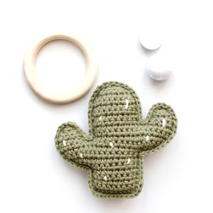 May include: A green crocheted cactus toy with white stitching. The cactus is on a white background with a wooden ring and two white beads.