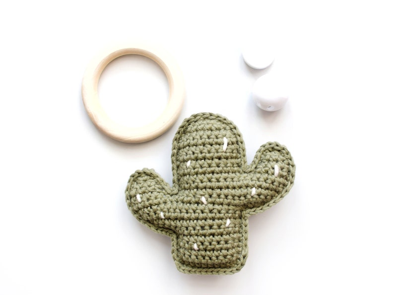 May include: A green crocheted cactus toy with white stitching. The cactus is on a white background with a wooden ring and two white beads.