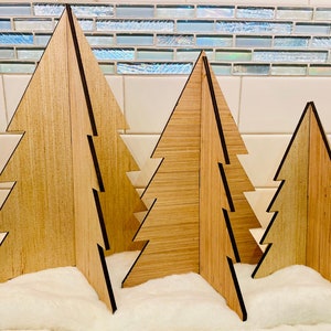 Pode incluir: Três árvores de Natal de madeira de tamanhos diferentes, em pé sobre uma superfície branca e fofa. As árvores são feitas de madeira marrom claro com bordas pretas e têm um design em camadas e em degraus.