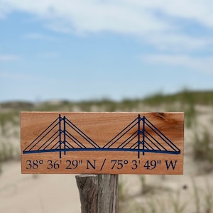 Puede incluir: Un letrero de madera con una silueta azul de un puente colgante y las coordenadas "38° 36' 29" N / 75° 3' 49" W" impresas en él.
