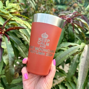 以下が含まれることがあります： 銀色の蓋が付いたさび色のステンレス製のタンブラー。タンブラーには「Keep Calm and Eat Mooncake Autumn 2024」という文字が刻まれています。