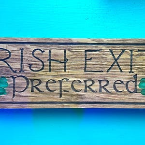 K&ouml;nnte beinhalten: Rechteckiges Holzschild mit dem Text "IRISH EXIT Preferred" eingeritzt. Das Schild hat gr&uuml;ne Kleeblatt-Details und einen dunklen Rahmen. Der Hintergrund ist in einem hellen T&uuml;rkis gehalten.