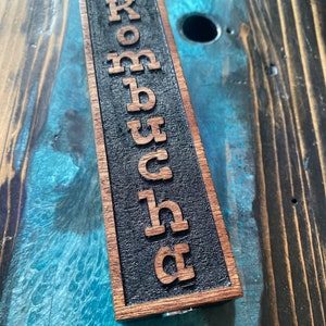 May include: A wooden sign with the word "Kombucha" carved into it. The sign is dark brown with black lettering and is sitting on a wooden surface.
