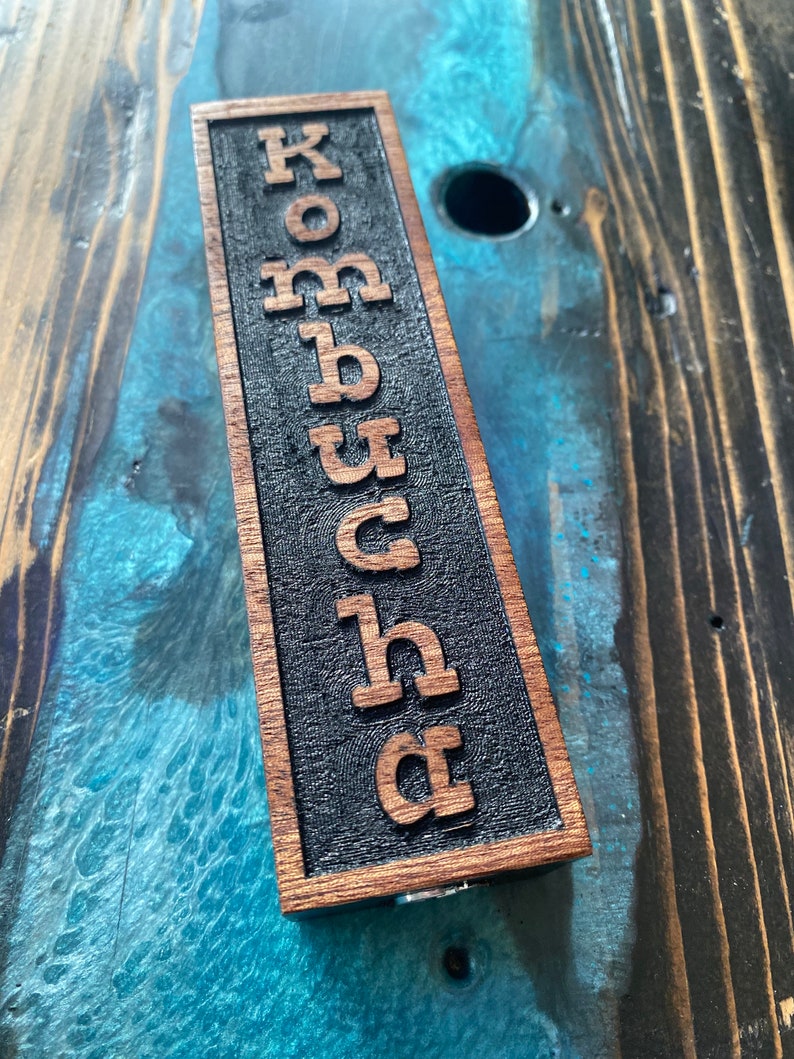 May include: A wooden sign with the word "Kombucha" carved into it. The sign is dark brown with black lettering and is sitting on a wooden surface.