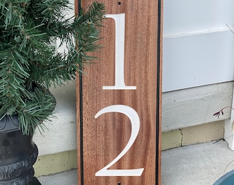 Puede incluir: Un letrero de número de casa de madera con borde negro. Los números "12" están tallados en la madera y pintados de blanco.