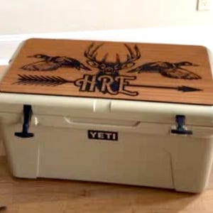 Puede incluir: Nevera beige con tapa de madera con un ciervo, patos y las letras "HRE" con una flecha. La nevera tiene cierres negros y la marca "YETI" en la parte delantera.