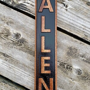 May include: A wooden sign with the name "ALLEN" carved into it. The letters are a light brown color and are set against a black background. The sign is framed in a dark brown wood.