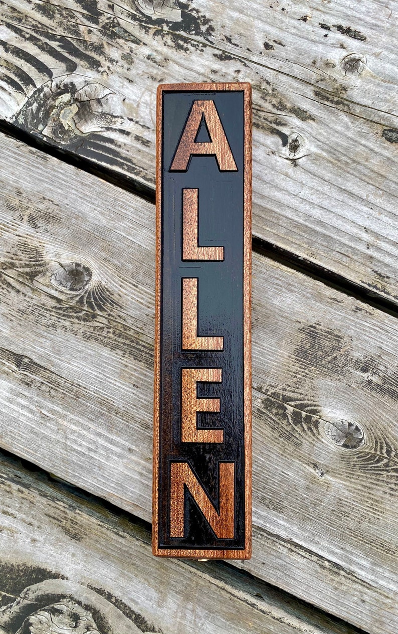 May include: A wooden sign with the name "ALLEN" carved into it. The letters are a light brown color and are set against a black background. The sign is framed in a dark brown wood.