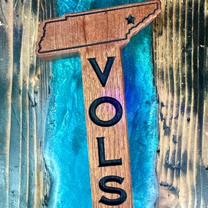May include: Wooden sign shaped like the state of Tennessee with the word "VOLS" carved into it. The sign is painted brown and has a star in the upper right corner.