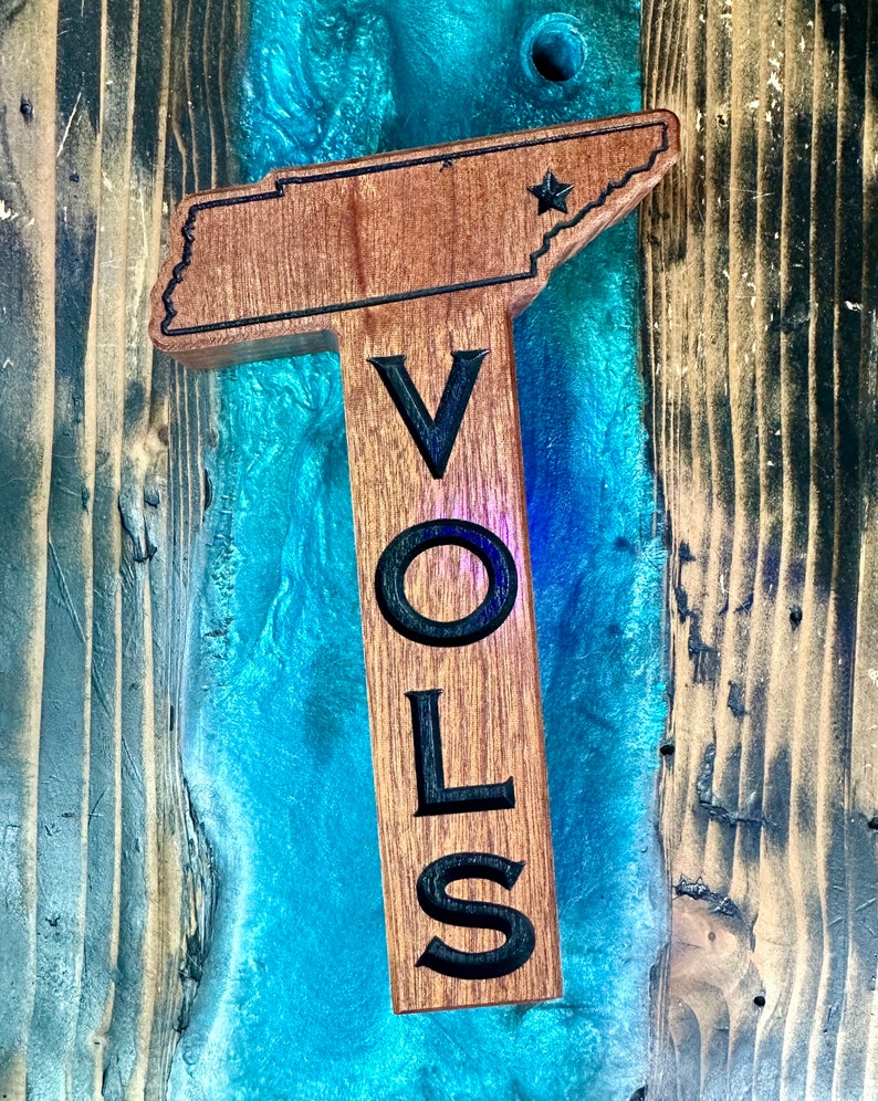 May include: Wooden sign shaped like the state of Tennessee with the word "VOLS" carved into it. The sign is painted brown and has a star in the upper right corner.