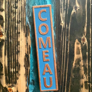 May include: A wooden sign with the word "COMEAU" painted in blue letters on a blue background.