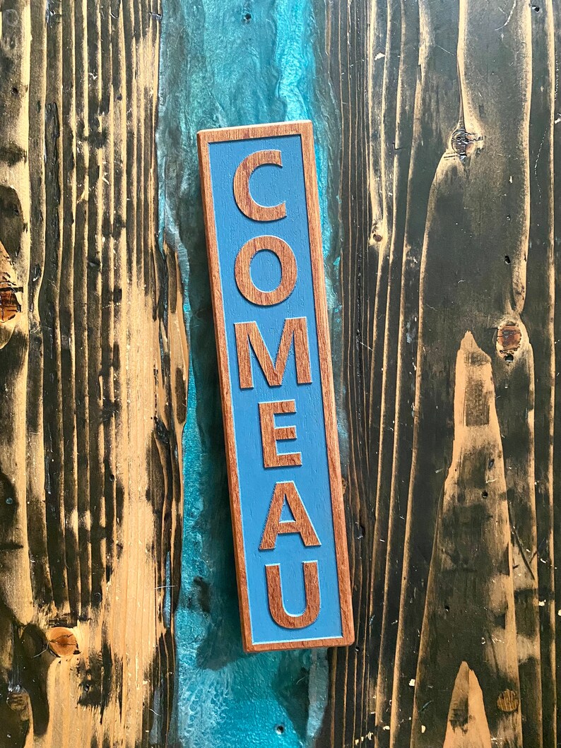 May include: A wooden sign with the word "COMEAU" painted in blue letters on a blue background.