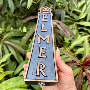 May include: A wooden beer tap handle shaped like a lighthouse with the word "ELMER" carved into it in gold lettering on a dark blue background.
