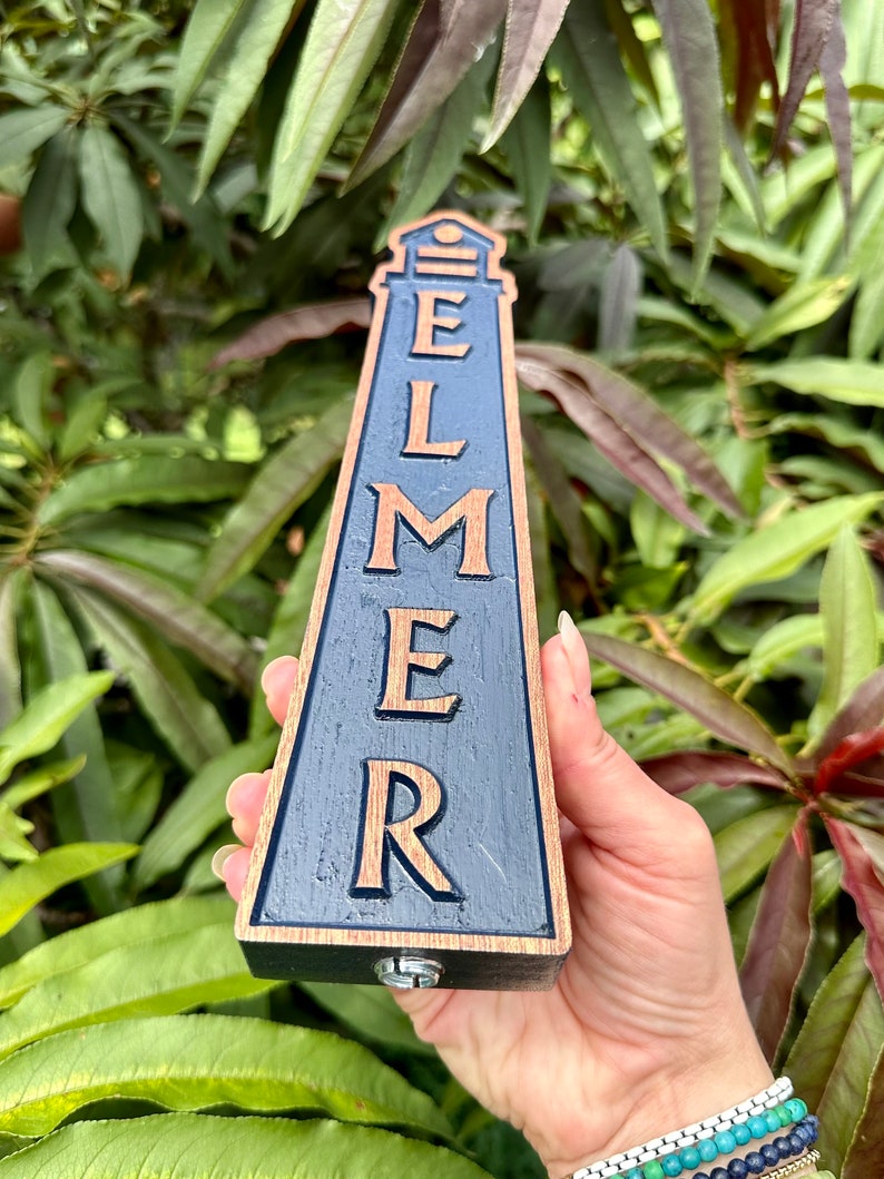 May include: A wooden beer tap handle shaped like a lighthouse with the word "ELMER" carved into it in gold lettering on a dark blue background.
