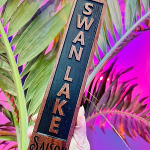 May include: A wooden tap handle with the words "SWAN LAKE SAISON" engraved on it. The handle is dark brown with black lettering.