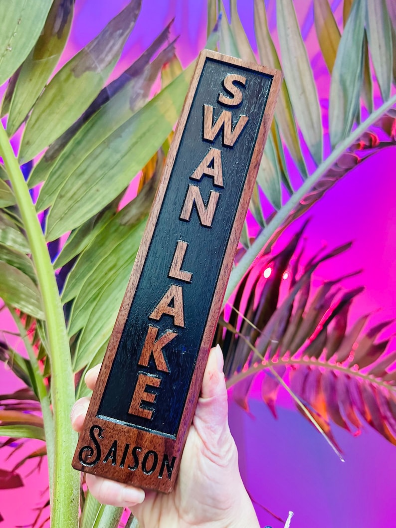 May include: A wooden tap handle with the words "SWAN LAKE SAISON" engraved on it. The handle is dark brown with black lettering.