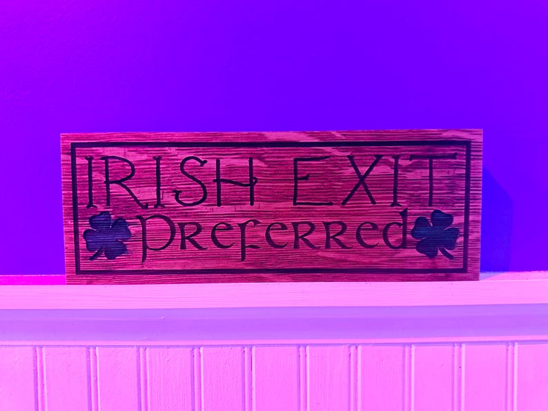 K&ouml;nnte beinhalten: Rechteckiges Holzschild mit den Worten "IRISH EXIT PREFERRED" in schwarzer Schrift. Das Schild hat einen dunklen Rand und zeigt auf jeder Seite ein Kleeblatt. Das Holz hat eine rotbraune Beize.