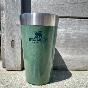 以下が含まれることがあります： ステンレス製の蓋が付いた緑色のスタンレーの断熱タンブラー。タンブラーには、翼のあるクマのロゴと「スタンレー1913年以来」の文字が書かれています。