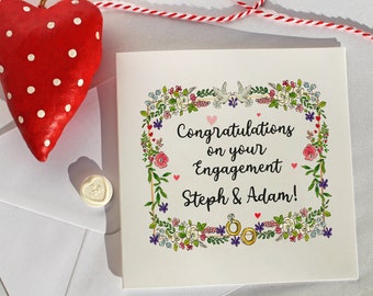 May include: An engagement card with a floral border, featuring the text "Congratulations on your Engagement Steph & Adam!". A red heart with white spots hangs above the card. A small white candy with "Love" text is also present.