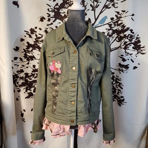 May include: An olive green denim jacket with gold buttons, featuring patches of camouflage and pink fabric. The jacket has a pink ruffled hem and cuffs, and decorative patches on the front.