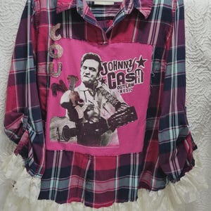 Puede incluir: Una camisa de franela patchwork con un estampado de cuadros rosas y azules. La camisa presenta un panel rosa con un gráfico de Johnny Cash y las palabras "Johnny Cash Outlaw Music". El dobladillo inferior está adornado con encaje blanco.