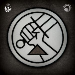 Puede incluir: Parche blanco bordado con un contorno negro de un puño sosteniendo un cuchillo, un triángulo y el texto "SKELETON CREW STUDIO, LLC".