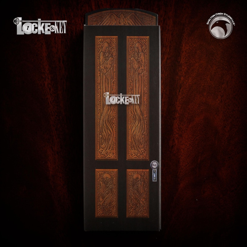 Pu&ograve; includere: Una scatola nera con un design a forma di porta in legno. La porta ha un design intagliato e una maniglia argentata. La scatola ha la scritta "Locke & Key" stampata su di essa.