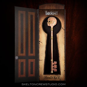 Puede incluir: Una llave de color dorado con un diseño de cabeza humana, en una caja con las palabras "Locke & Key" y "Head Key" en ella.