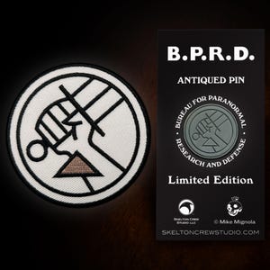 Puede incluir: Parche bordado en blanco y negro con un diseño de puño estilizado y un triángulo marrón. Junto a él, una tarjeta negra con el texto "B.P.R.D. ANTIQUED PIN" y "BUREAU FOR PARANORMAL RESEARCH AND DEFENSE".