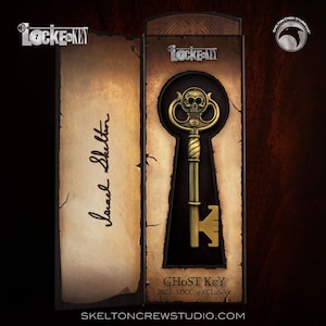 Puede incluir: Una llave dorada con un diseño de calavera en la parte superior, en una caja con el texto "Locke & Key" y "Ghost Key 2022 SDCC Exclusive".