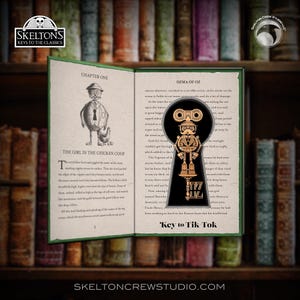 May include: An open book with a key-shaped pendant featuring a robot design. The pendant is gold-colored and has the text "Key to Tik Tok". The book is titled "Ozma of Oz" and "The Girl in the Chicken Coop". The background is a bookshelf.