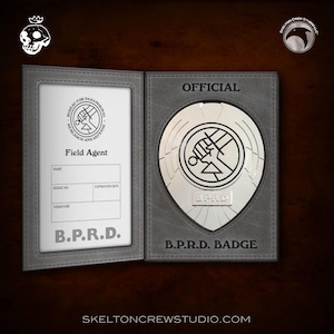 May include: A silver B.P.R.D. badge featuring a black fist and a circle with a line through it. The badge is housed in a grey leather case with the words "OFFICIAL" and "B.P.R.D. BADGE" printed on it.