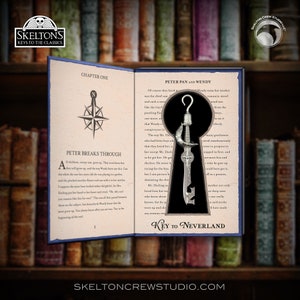 Puede incluir: Una llave plateada con diseño de espada y un gancho en la parte superior. La llave está delante de un libro abierto con el texto "Peter Pan y Wendy" y "Llave a Nunca Jamás".