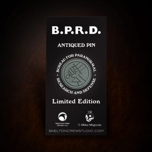 Puede incluir: Una tarjeta rectangular negra con texto blanco que dice "B.P.R.D. ANTIQUED PIN" encima de un emblema circular. El emblema tiene las palabras "BUREAU FOR PARANORMAL RESEARCH AND DEFENSE" alrededor. Debajo está "Limited Edition".