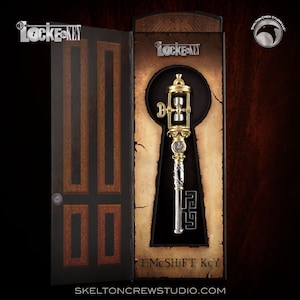 Può includere: Una chiave dorata e argentata con un quadrante dell'orologio e una clessidra nella parte superiore. La chiave è in una scatola con le parole "TimeShift Key" e "Locke & Key" su di essa.