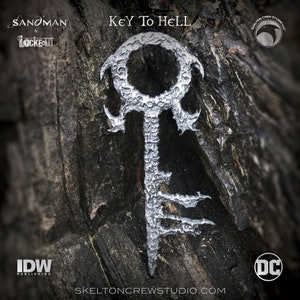 Può includere: Una chiave d'argento con un design dettagliato, etichettata "Key to Hell" dalla serie di fumetti Sandman & Locke & Key. La chiave è su uno sfondo di legno scuro con i loghi IDW Publishing e DC Comics.