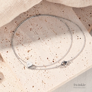 K&ouml;nnte beinhalten: Eine silberne Kettenkette mit einem kleinen Herz-Anh&auml;nger mit einem Buchstaben 'K' auf einer hellbeige strukturierten Oberfl&auml;che. Die Kette hat einen Karabinerverschluss. Der Text 'Twinkle Personalized' ist am unteren Rand des Bildes sichtbar.