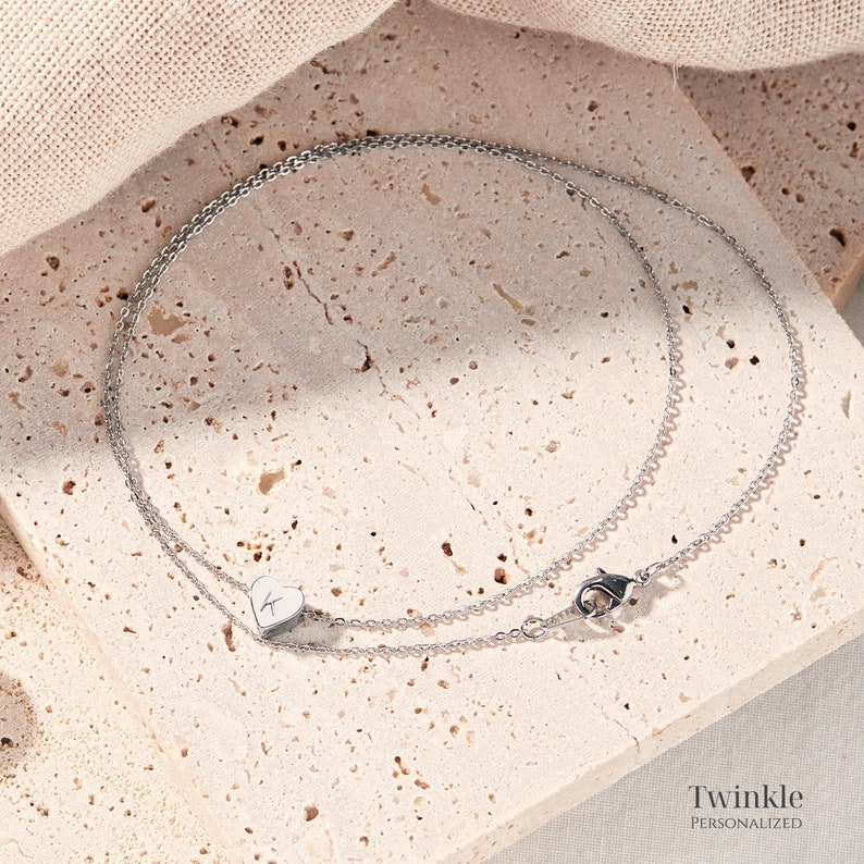 K&ouml;nnte beinhalten: Eine silberne Kettenkette mit einem kleinen Herz-Anh&auml;nger mit einem Buchstaben 'K' auf einer hellbeige strukturierten Oberfl&auml;che. Die Kette hat einen Karabinerverschluss. Der Text 'Twinkle Personalized' ist am unteren Rand des Bildes sichtbar.